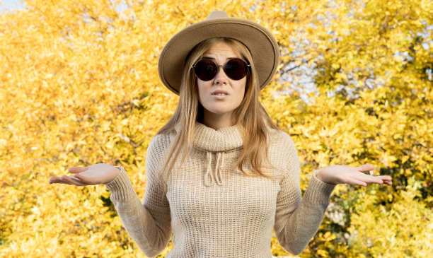 戴着帽子的惊讶女子站在秋天的公园，黄色的树叶背景。穿着暖和毛衣的女孩在散步。温暖的阳光明媚的天气。秋天树叶图片下载