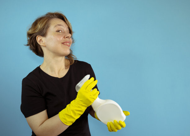 快乐的清洁妇女在黑色t恤拿着白色喷枪在孤立的蓝色背景。戴着黄色橡胶清洁手套的女人。portrai图片下载