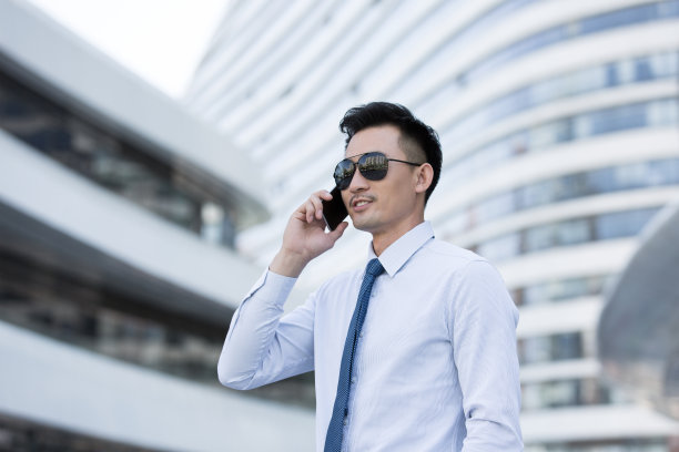中年商人戴着墨镜，穿着职业装，在办公楼外用智能手机打电话——照片图片下载
