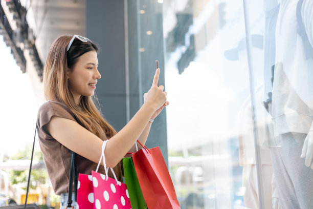 美丽的女人拿着购物袋用手机在时装店的橱窗外拍照。时尚女性在服装店附近拿着智能手机。女性购物生活时尚图片下载