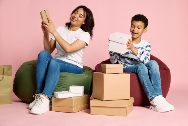 画像的女人坐在十几岁的男孩和打开盒子购物后孤立的粉红色工作室背景图片下载