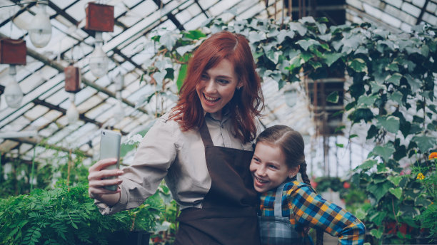 漂亮的温室女主人和她可爱的女儿在果园里用智能手机自拍，摆姿势，微笑。人与技术的概念。图片下载