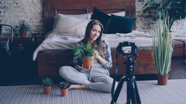 穿着休闲装、面带微笑的年轻博主正手持鲜花，用三脚架上的相机为在线视频博客录制关于室内植物的视频博客。图片下载