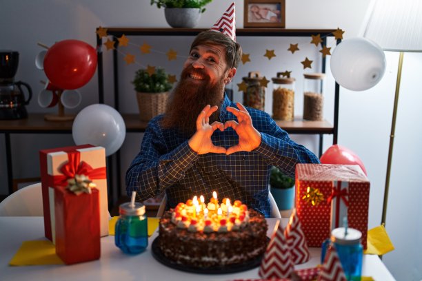 留着长胡子的白种人男子手持大巧克力蛋糕庆祝生日，用手展示心形符号和形状，面带爱意地微笑。浪漫的概念。图片下载