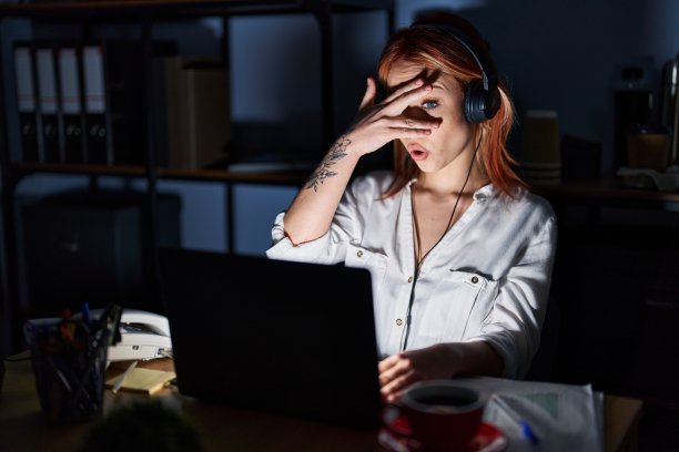 晚上在办公室工作的年轻白人女性用手捂着脸和眼睛，用手指向外张望，露出尴尬的表情。图片下载