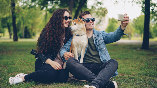 英俊的年轻人正在与他漂亮的妻子和可爱的狗狗自拍，他们都戴着墨镜。这家伙拿着智能手机拍照摆姿势。图片下载