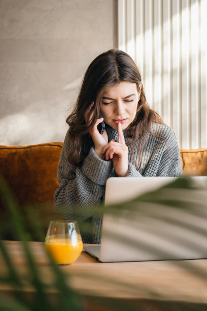 迷人的年轻女子一边打电话一边用笔记本电脑工作。图片下载