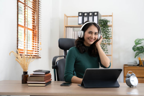 画像微笑的亚洲女性戴着现代耳机远程工作，通过视频通话应用程序与同事沟通。图片下载