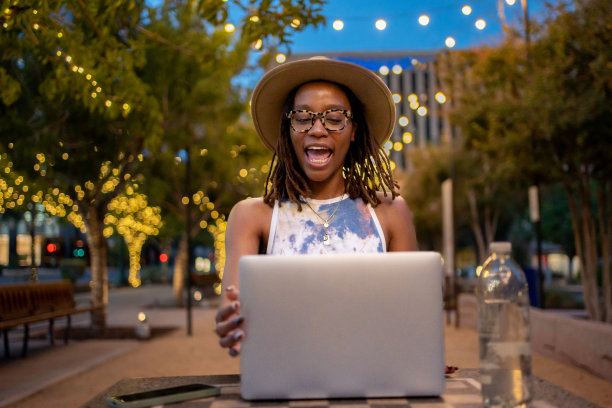 在德克萨斯州埃尔帕索的一个公园里，20多岁的美丽的非裔美国妇女戴着帽子，在装饰性的灯光下用笔记本电脑进行电话会议图片下载