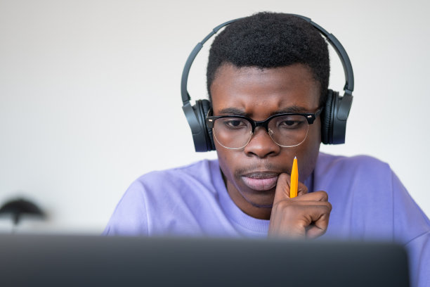 非洲年轻人戴着耳机用笔记本电脑工作，看起来沉思图片下载