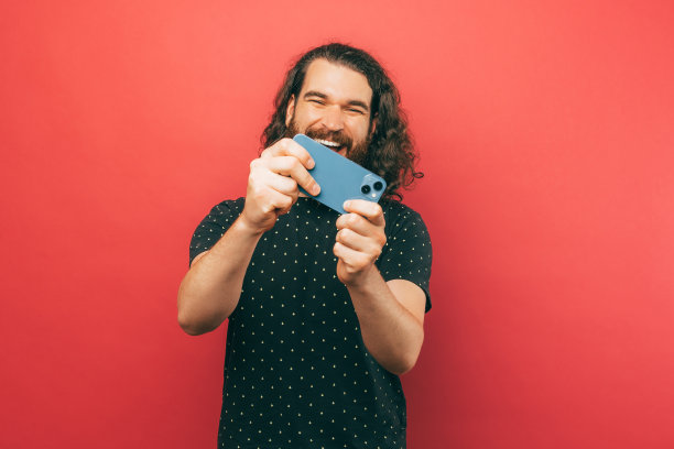 照片上，年轻的蓄着胡子、留着长发的男子在粉色背景下玩着智能手机上的网络游戏。图片下载