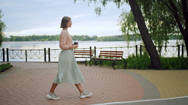 穿着粉色衬衫和绿色长裙的年轻女性端着一杯咖啡走在码头上图片下载