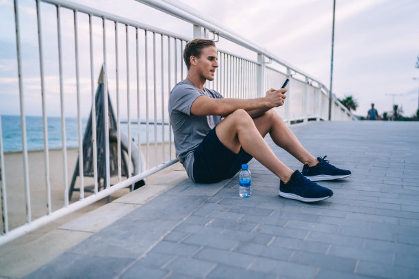 年轻的白人运动员坐在户外休息后，训练检查邮件和发送短信在手机上，苗条的潮人保持健康的生活方式使用智能手机上的训练应用程序图片下载