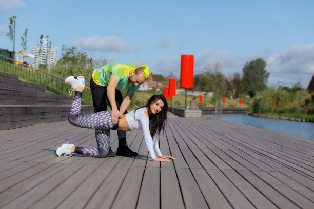 一名男教练帮助一名身穿运动服的褐发女子在街头操场上进行腿摆动练习图片下载