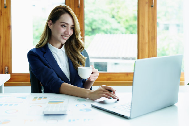 画像的亚洲商业女性喝咖啡，工作与电脑和财务报表文件在她的办公桌上图片下载