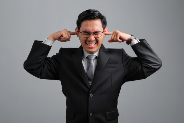 穿着正装的年轻商人心烦意乱，用手指堵住耳朵，避免在灰色背景中隔绝不愉快的声音图片下载