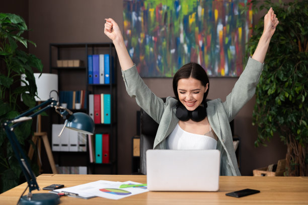快乐兴奋的年轻女子举手，坐在家里的办公桌前工作，从远处在线学习，戴着耳机和智能西装，谈话面试后胜利图片下载
