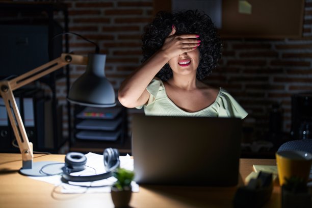 年轻的黑发女人和卷发在办公室工作在晚上微笑和大笑的手在脸上掩盖眼睛惊讶。盲目的概念。图片下载