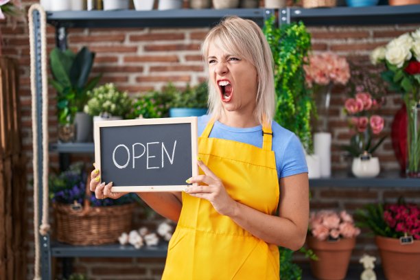 年轻的白人妇女在花店工作，拿着开着的牌子生气和疯狂的尖叫，沮丧和愤怒，叫喊着愤怒。愤怒和攻击性的概念。图片下载