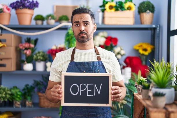 留胡子的西班牙人在花店工作，拿着打开的牌子，怀疑和紧张，因为问题皱着眉头心烦意乱。消极的人。图片下载