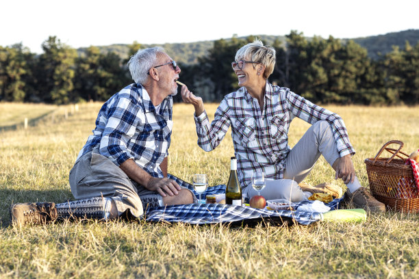 成熟的情侣坐在大自然的毯子上野餐。她用一块食物喂老人，微笑着图片下载