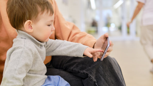 拿着手机的母亲和婴儿在诊所等待儿科医生。父母带着手里拿着智能手机的小男孩。孩子是一个一岁零三个月的男孩图片下载