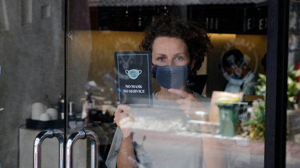 一名妇女指着餐厅门口的无口罩无服务海报，这是全球大流行期间的冠状病毒防护措施。限制大流行病的世界创新和变化。图片下载