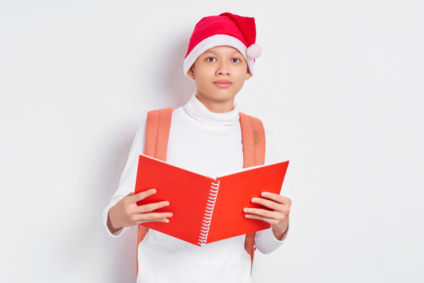 微笑英俊的年轻亚洲学生，戴着圣诞帽和休闲服，背着双肩包，拿着白色背景的书图片下载