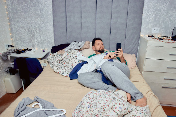 一个手里拿着电话的男人躺在卧室的床上，衣服散乱，卧室里一片狼藉。图片下载