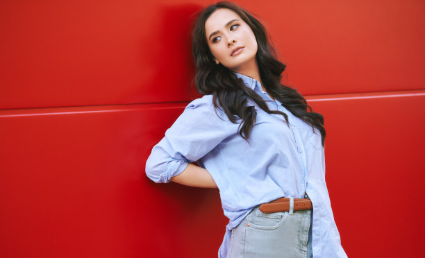 户外时尚肖像漂亮的年轻女子靠在红色的墙壁，穿着蓝色的衬衫，自然的妆容和波浪发型图片下载