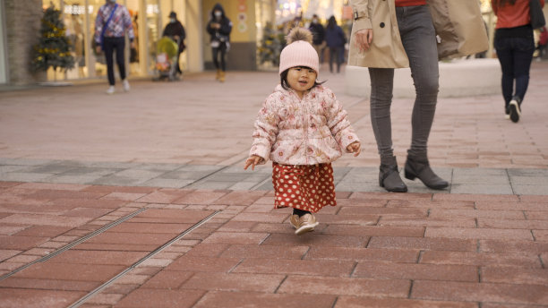 全长搞笑的亚洲小女孩在圣诞气氛装饰的购物街上玩得很开心。一个母亲走在后面照顾她的短镜头图片下载
