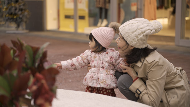 后视图，亚洲男孩与女婴和她的母亲分享玩具，女婴和她的母亲看着人们在下雪的城市公园里玩耍。童年的友谊概念图片下载