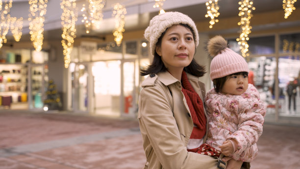 一个可爱的亚洲小女孩正指着上面闪闪发光的圣诞彩灯，而她的妈妈在寒假期间走在灯火通明的购物街上图片下载