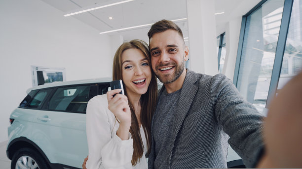 迷人的年轻情侣在现代汽车展厅用车钥匙自拍。他们看着镜头，亲吻，微笑。图片下载