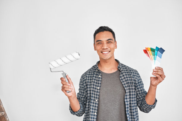 一个年轻的亚洲人拿着油漆辊和色板对着空白的墙壁。图片下载