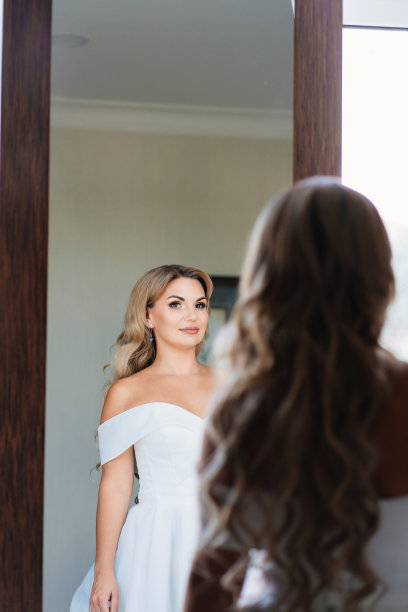 美丽的金发新娘穿着白色的裙子看着镜子里的倒影。图片下载