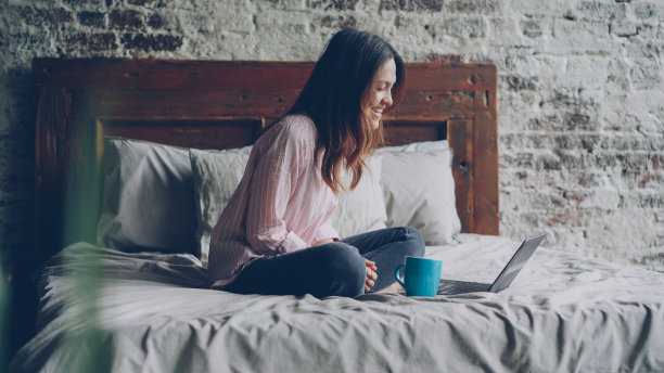 穿着休闲装的漂亮年轻女子坐在卧室的双人床上，用笔记本电脑和朋友聊天。技术与传播概念图片下载