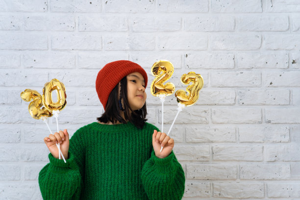 戴着红帽子和绿色毛衣的亚洲可爱女孩拿着印有数字2023的铝箔气球。图片下载