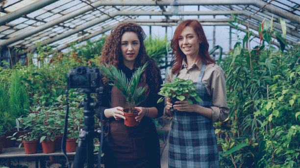 面带微笑的女博主和穿着围裙的园丁手持鲜花交谈，用三脚架上的相机为在线视频博客录制关于园艺的视频博客。图片下载