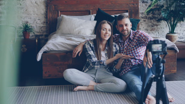一对可爱的情侣正在用相机拍摄社交媒体视频，他们穿着休闲装坐在阁楼式卧室的地板上与粉丝交谈。图片下载