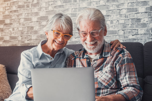 可爱的老夫妇坐在沙发上一起用笔记本电脑购物和上网。两个戴着眼镜的成年人在客厅享受科技。老人们在爱情中欢笑的画像。图片下载