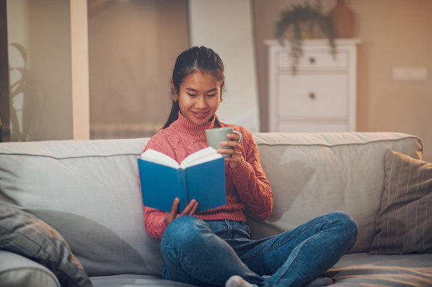 亚洲女人坐在家里的沙发上，一边看书一边喝咖啡图片下载