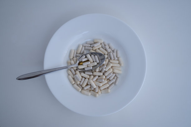 白色的盘子里装着米色的药片和药品，白色背景上有一个金属勺子。比喻疾病的治疗。盘子里的维生素可以预防和治疗疾病。图片下载