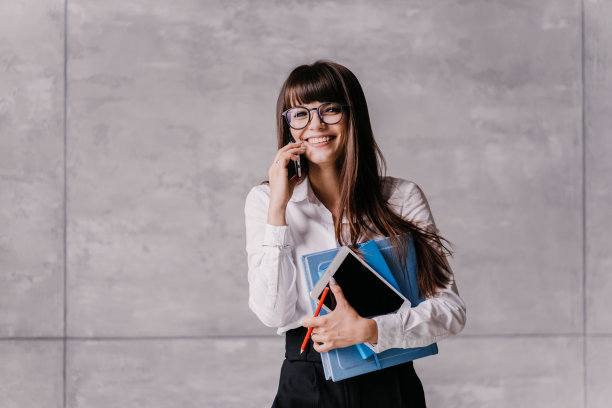 快乐的黑发女人，戴着眼镜，白衬衫和黑裤子，拿着平板电脑，日记，露齿微笑，看向一边，说着电话。笑脸学生女孩准备上课。教育和商业图片下载