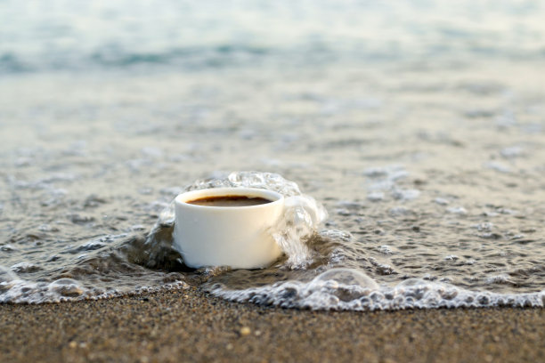 咖啡杯顶着海水，海浪滔天。杯子放在沙滩上或大的海滨岩石上。早晨的假期，水打杯咖啡图片下载