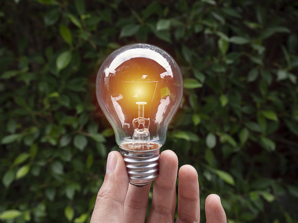 女人手里拿着灯泡，橙色的灯在绿色的自然背景下发光。图片下载