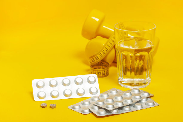 银盘子旁边放着没有包装的片剂和药片，背景是一杯饮用水和运动设备:哑铃，卷尺。副本的空间。图片下载