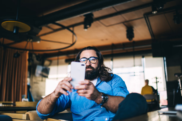 40多岁的留着胡子的白人男子在空闲时间坐在咖啡馆里用智能手机看视频，英俊的成熟男子拿着手机阅读信息和网上聊天图片下载