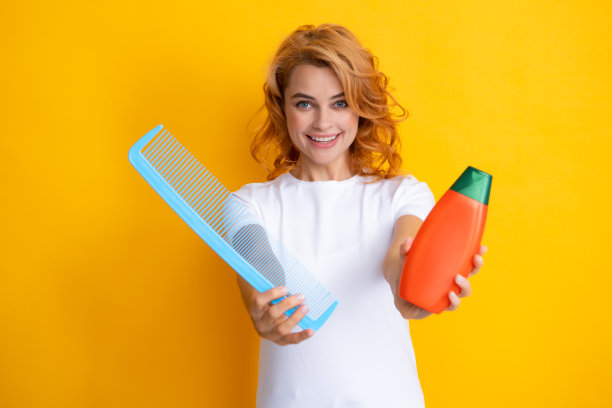 有趣的肖像红发女人梳，孤立在黄色背景。妇女拿着一瓶洗发水和护发素。图片下载