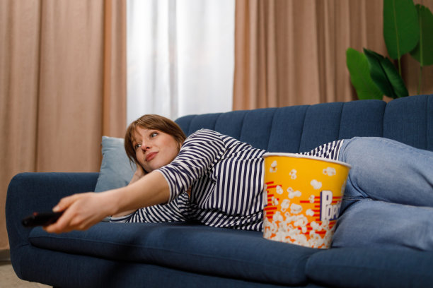 快乐的年轻女子与遥控器放松在家里的沙发上，看电视和吃爆米花图片下载
